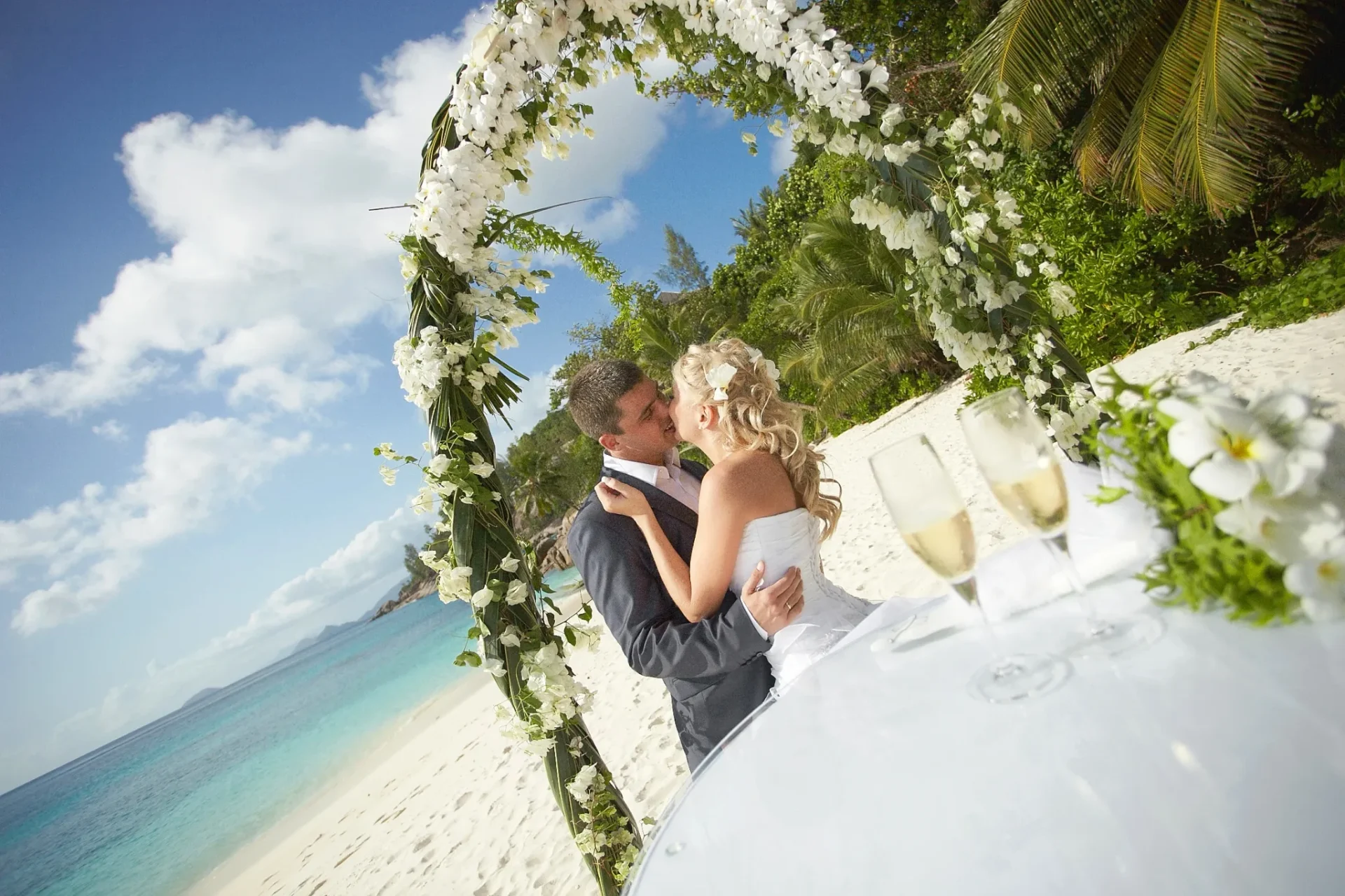 Couple kissing under a white floral wedding arch on a St. Thomas beach with turquoise water and champagne glasses in the foreground.